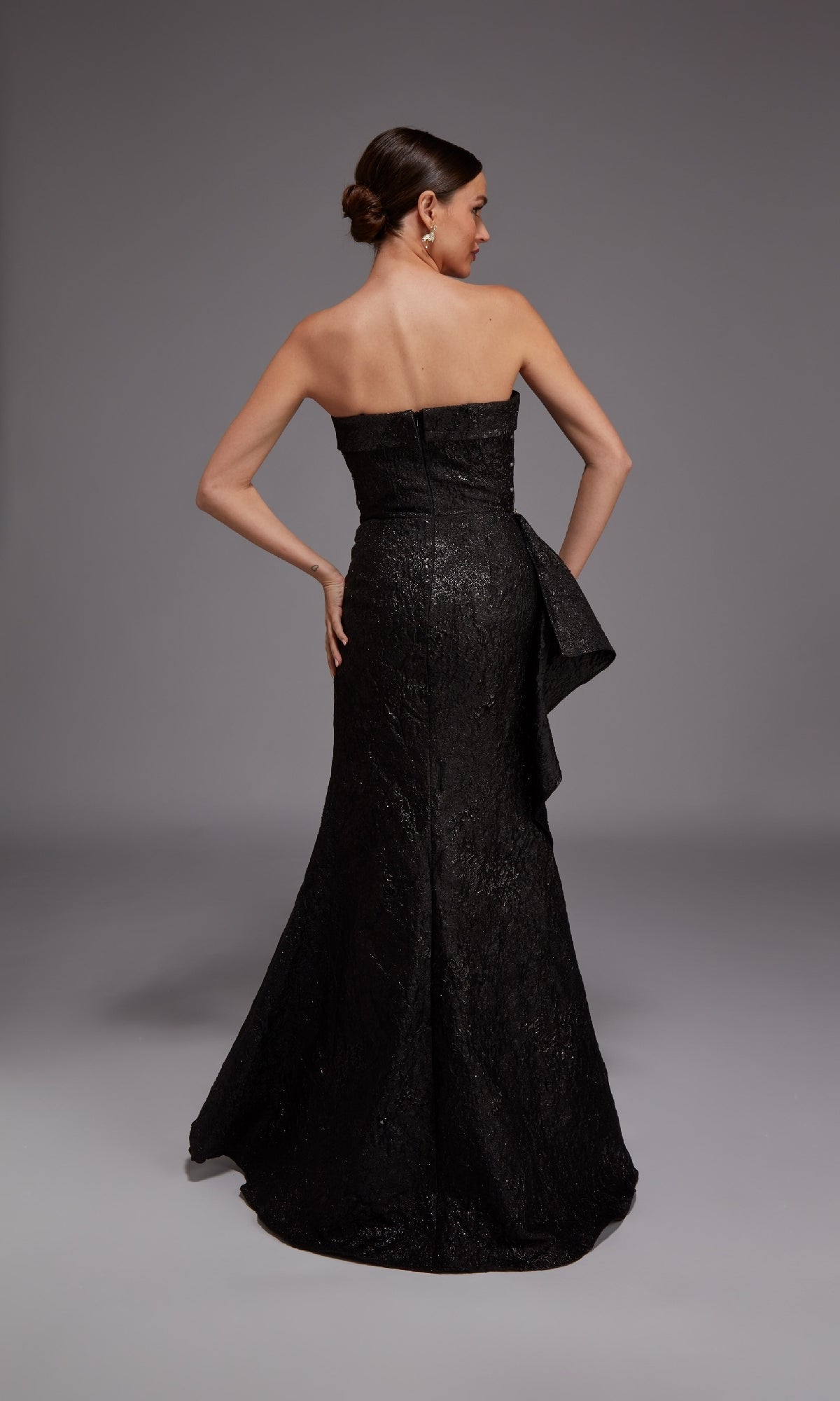 A woman in the Alyce 27818 Long Formal Dress, featuring a strapless jacquard design with a fitted bodice and flared skirt, stands facing away with hand on hip against a plain gray background—elegance from every angle.